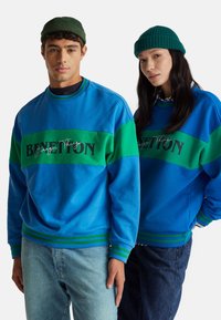 Felpa blu con striscia verde, con scritta nera "BENETTON". Polsini e orlo a coste con strisce verdi a contrasto. Indossata con jeans casual.