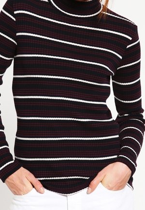Person iført en sort, hvid og vinrød vandret striped ribstrikket langærmet rullekrave sweater med hænderne i lommerne på hvide bukser.
