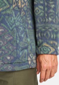 Fleece jacket with a blue, green, and yellow geometric pattern, featuring a side pocket and rolled cuff. Soft texture, relaxed fit.
