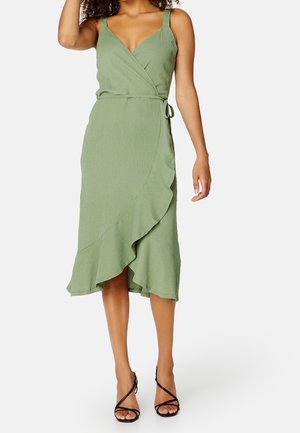 Kobieta nosząca bezrękawową, jasnoliściastą sukienkę kopertową z falbanką u dołu i cienkim paskiem do wiązania, w połączeniu z czarnymi sandałami na wysokim obcasie z paskami.