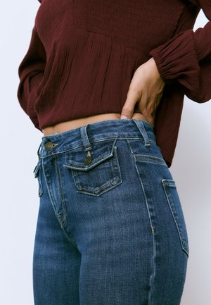 Dark blue denim jeans with a fitted shape, featuring two front pockets and two back pockets. The waistband includes a brass button detail.