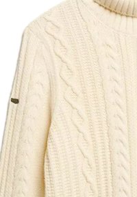 Cremefarvet kabelstrikket højhalset sweater med strukturerede fletmønstre og et lille sort mærke på venstre ærme.