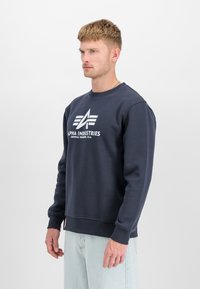 Tumši zilā džempera krekls, kas izgatavots no maiga kokvilnas maisījuma, ar apaļu izgriezumu, ribotām piedurknēm un baltu logotipu uz priekšpuses.