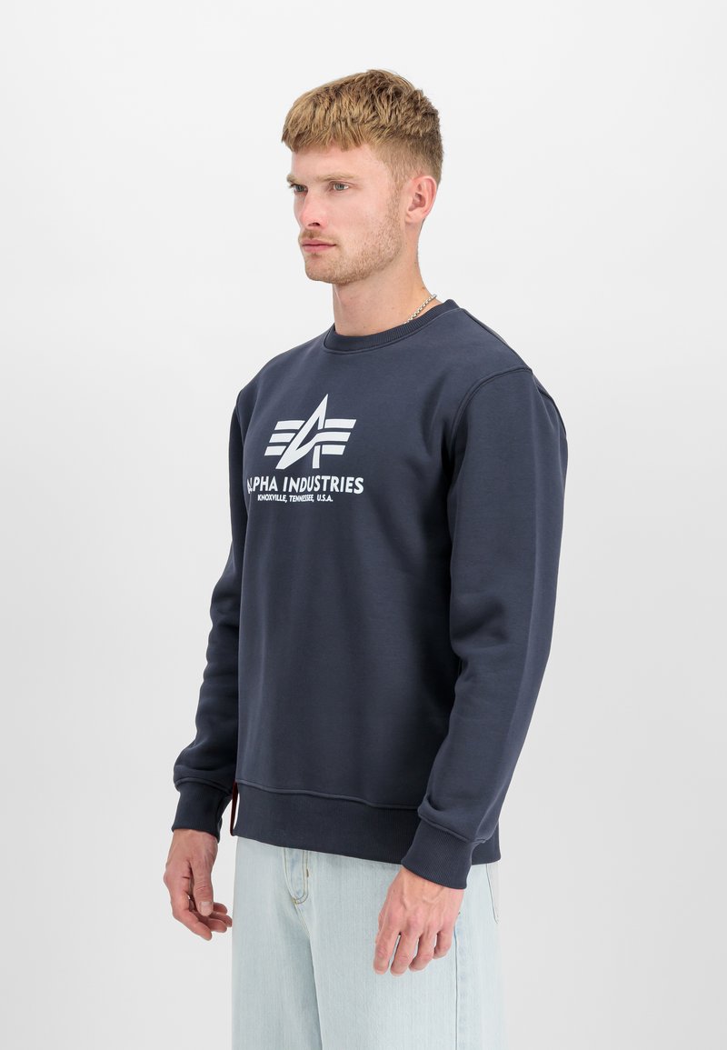 Tumši zilā džempera krekls, kas izgatavots no maiga kokvilnas maisījuma, ar apaļu izgriezumu, ribotām piedurknēm un baltu logotipu uz priekšpuses.