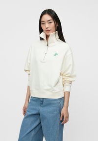 Kreminė zip-up džemperis su aukšta apykakle ir žaliai logotipu. Minkšta medžiaga, laisvas kirpimas ir riboti rankogaliai. Derinamas su šviesiai mėlynomis džinsinėmis kelnėmis.