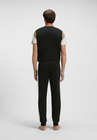 Man staat blootsvoets met zijn rug naar ons gekeerd, draagt zwarte broek met witte zijstrepen en een zwart, wit en tan shirt met korte mouwen.