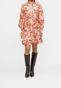 Femme portant une robe rose à fleurs à manches longues avec une ceinture à la taille et des bottes en cuir noires montant jusqu’aux genoux, debout devant un fond uni.