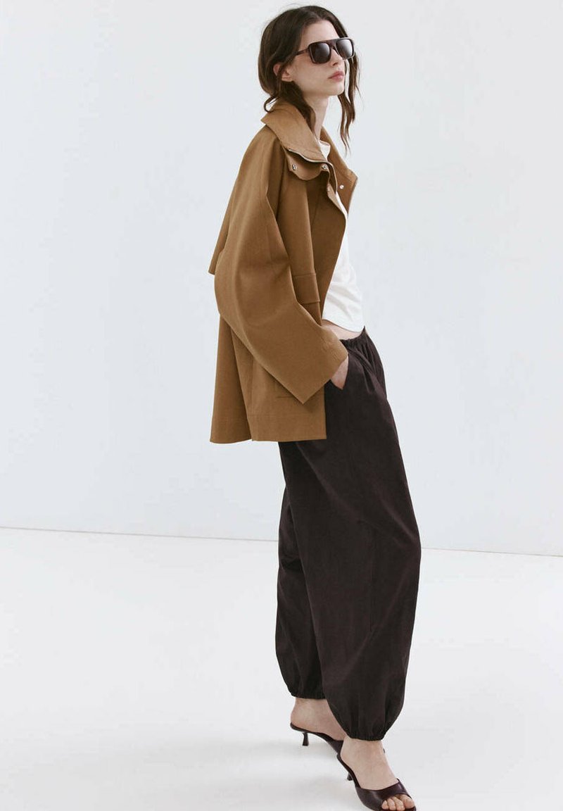 Femme portant une veste marron oversize, un haut blanc, un pantalon noir ample, des sandales à talons noires et des lunettes de soleil foncées sur un fond clair uni.