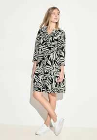 Schwarzes Kleid mit einem Blattdruck in Hellgrün, kragenförmiger Ausschnitt, Dreiviertelärmel und Taschen. Kombiniert mit weißen Sneakers.