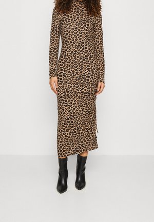 Vrouw draagt een jurk met lange mouwen en een luipaardprint tot op de enkel met zwarte puntige enkellaarzen tegen een effen achtergrond.