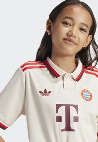 Maillot de football beige avec des accents rouges, un col blanc, un design à trois rayures rouges sur les épaules, un logo à l'avant en bordeaux, et un emblème de club sur la poitrine.