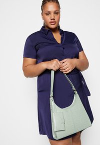 Borsa a tracolla verde con texture a forma di coccodrillo, dotata di una tasca frontale. Indossata con un vestito camicia blu navy con bottoni.