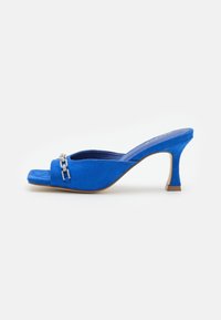 Mule de ante azul con punta abierta y un pequeño tacón de aguja, que presenta un detalle de cadena plateada en la parte delantera. Textura suave y diseño elegante.