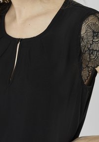 Black blouse featuring a rounded neckline and a keyhole opening, with sheer lace sleeves and a soft, textured fabric.