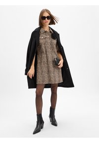 Leopard-print jurk met een knoopsluiting kraag, gecombineerd met een zwarte jas. Model draagt zwarte zonnebril en enkellaarsjes. Zwarte clutch in de hand.