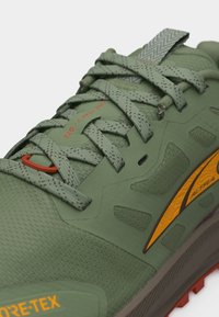 Πράσινο παπούτσι για τρέξιμο σε μονοπάτια με κορδόνια με σχέδια, πορτοκαλί λεπτομέρειες και σήμανση Gore-Tex στην περιοχή των δακτύλων.