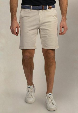 Mann trägt beige Shorts mit umgekrempeltem Saum, einen blauen gewebten Gürtel und weiße Sneakers, steht vor einem einfarbigen Hintergrund.