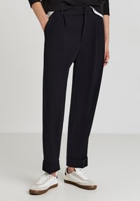 Schwarz tapered Hose mit glatter Textur, einem gefütterten Bund und hochgerollten Säumen, kombiniert mit weißen und grauen Sneakers.