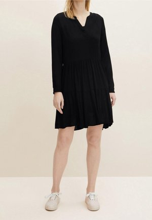 Femme portant une robe noire ample à manches longues à étages et des baskets blanches, debout devant un fond beige clair uni.