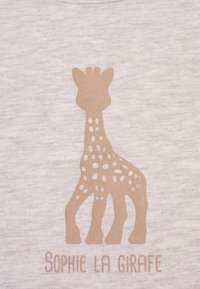 T-shirt in cotone beige con una silhouette stilizzata di giraffa con macchie bianche e il testo "Sophie La Girafe" in basso di colore abbinato.