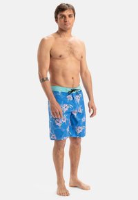 Blå badbyxor med ett blommönster i rosa och grönt. Har en ljusgrön midja och svart snörning, tillverkade av lättviktsmaterial.