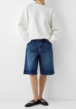 Persoon die een oversized wit sweatshirt draagt, blauwe knielange spijkershorts en zwarte geweven platte schoenen, staand met handen in de zakken.