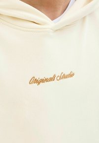 Pogled od blizu na kremasti duksericu s vezenim natpisom "Originals Studio" smešnim smešnim slovima u smešnoj boji na grudima.