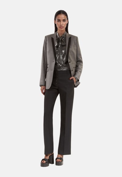Grauer, maßgeschneiderter Blazer mit schwarzen Akzenten, glitzerndem Top und hoch taillierten schwarzen Hosen. Das Model trägt Plateauschuhe.