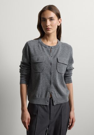 Donna che indossa un cardigan grigio abbottonato con due tasche sul petto e pantaloni grigio scuro, in piedi davanti a uno sfondo chiaro e uniforme.