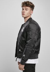 Črna bomber jakna z gladko, sijočo površino, z zadrgo spredaj, rebrastimi manšetami in logotipom na prsih ter rokavu.