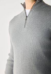 Suéter gris de canalé con media cremallera. La tela parece suave con un acabado texturizado, presentando un diseño ajustado y un cuello alzado.