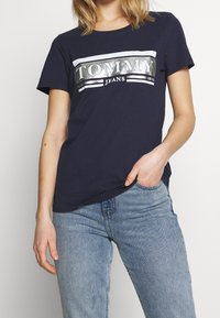 Námořnická modrá tričko vyrobené z bavlny, s reflexním stříbrným logem "TOMMY JEANS" napříč hrudníkem a bílými pruhy nad a pod ním.