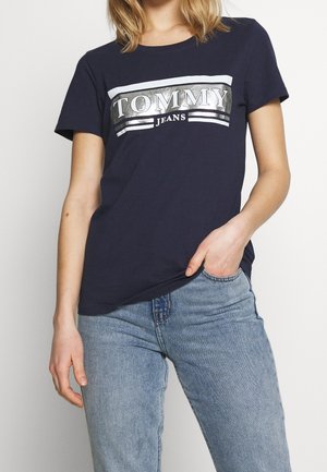 Camiseta azul marino de algodón, con un logo reflectante plateado "TOMMY JEANS" en el pecho y rayas blancas arriba y abajo.