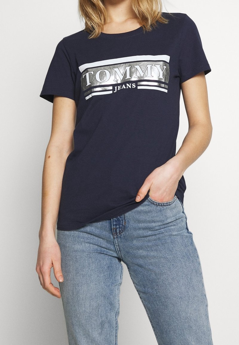 Námořnická modrá tričko vyrobené z bavlny, s reflexním stříbrným logem "TOMMY JEANS" napříč hrudníkem a bílými pruhy nad a pod ním.