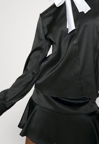 Blusa nera in satin con maniche lunghe e chiusura a bottoni, caratterizzata da una vestibilità ampia e un grande fiocco bianco al colletto.