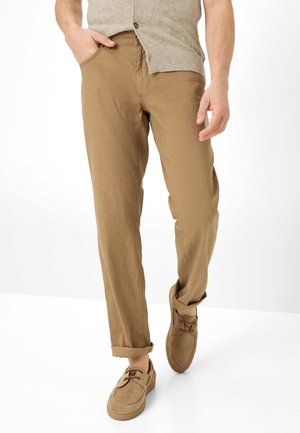 Uomo che indossa pantaloni marroni risvoltati, un gilet lavorato a maglia beige e scarpe in camoscio marroni abbinate, che cammina con una mano in tasca.