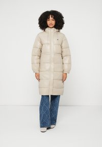 Casaco acolchoado bege claro com capuz, costuras visíveis e corte descontraído. Combinado com jeans azuis estampados com corte flare e botas de cor clara.