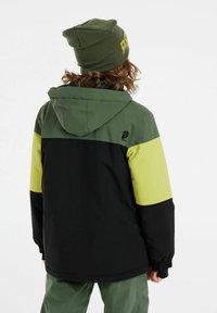 Veste d'hiver colorée en noir, vert et jaune. Dotée d'une capuche, de poignets élastiques et d'un logo sur l'épaule. Portée avec un bonnet assorti.