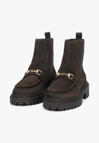 Brązowe zamszowe botki na kostkę z grubą, bieżnikowaną podeszwą, ozdobione dekoracyjnym złotym łańcuchem z przodu oraz kontrastowymi panelami po bokach.