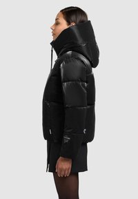 Črno puhovko z visokim ovratnikom, sijajnim teksturo, z zadrgo spredaj in stranskimi žepi. Ima šive in zaščitni znak na rokavu.