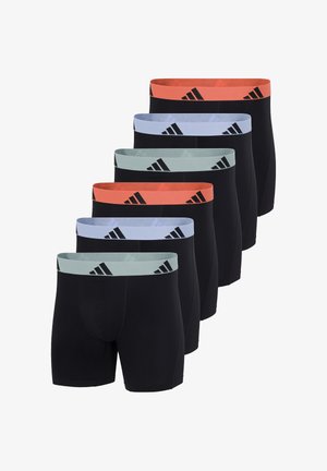 Athletischer Mann steht barfuß und trägt schwarze Boxer-Shorts mit einem orangefarbenen Bund, der schwarze Adidas-Logos zeigt.