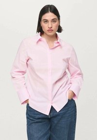 Cămașă roz deschis cu nasturi, cu rever clasic, mâneci lungi și croială lejeră. Purătată cu blugi de denim albastru. Textură fină a țesăturii.