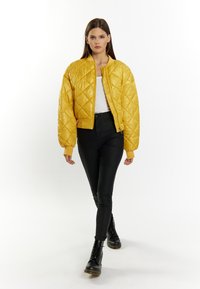 Giacca bomber trapuntata gialla con chiusura a zip, polsini a coste e colletto rialzato, abbinata a pantaloni skinny neri e stivali con lacci.