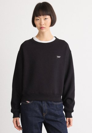 Black sweatshirts with a loose fit, crew neck, and ribbed cuffs. Features a small white logo on the chest and worn with dark denim jeans.