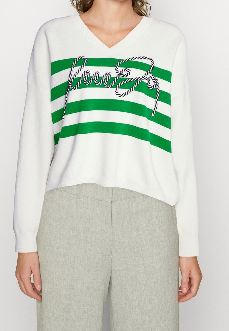 Pull blanc à col en V avec trois rayures horizontales vertes et une corde torsadée décorative formant le mot « Good » sur la poitrine.