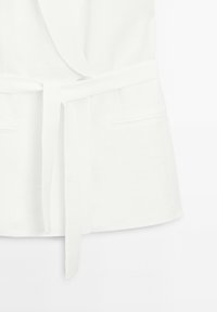 Valkoinen kietaisutakki vyöllä, jossa kaksi etutaskua ja sileä pinta. Yksinkertainen muotoilu, sopii kerrospukeutumiseen.