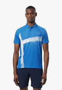 Sininen polo-paita, jossa on valkoinen roisketuloste. Paita sisältää kauluksen, kaksi nappia ja lyhyet hihat. Lacoste-logo vasemmalla rintakehällä.