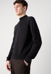 Schwarzer Rundhalsausschnitt-Pullover mit geripptem Kragen und Ärmeln, der eine glatte Textur aufweist. Modell trägt ihn mit dunkelbraunen Hosen und die Hände in den Taschen.