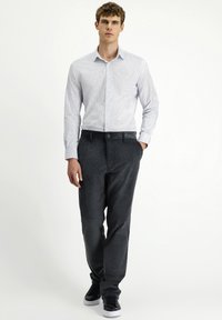 Overhemd met knoopsluiting in lichtblauw en witte strepen, gecombineerd met donker gestructureerde broek. Model draagt zwarte sneakers en heeft zijn handen in zijn zakken.