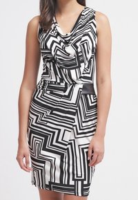 Vestido en blanco y negro con un patrón geométrico, escote drapeado y diseño ajustado, que presenta acentos sólidos en negro contrastante en la cintura.